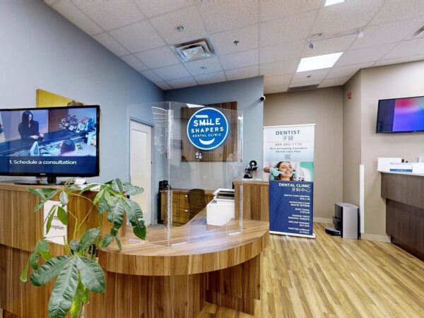 A modern dental office reception area featuring a wooden front desk, a television displaying content, a plant, and promotional materials on a stand. The walls are painted in neutral tones with blue accents, creating a welcoming atmosphere.