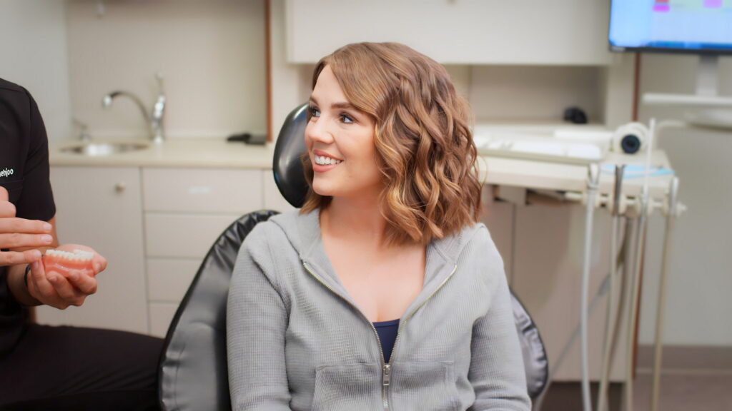 A smiling woman with wavy hair sits in a dental office, looking towards a healthcare professional who is holding a dental tool. The atmosphere is friendly and relaxed, suggesting a positive experience. The 123Dentist Smiley is present, adding a cheerful touch to the scene.