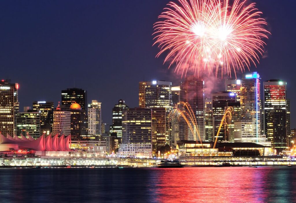 A vibrant city skyline at night, illuminated by colorful fireworks in the sky. Reflective water in the foreground adds to the festive atmosphere.
