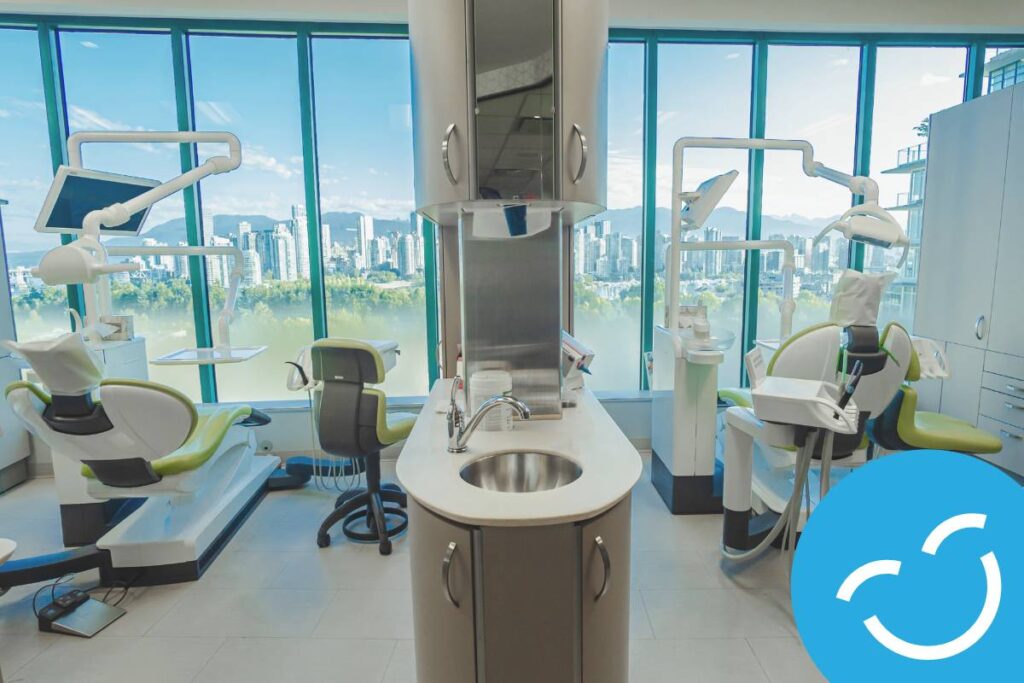 A modern dental clinic with large windows offering a city view. The image features dental chairs, equipment, and a central workstation with a sink, creating a clean and inviting environment for patients.