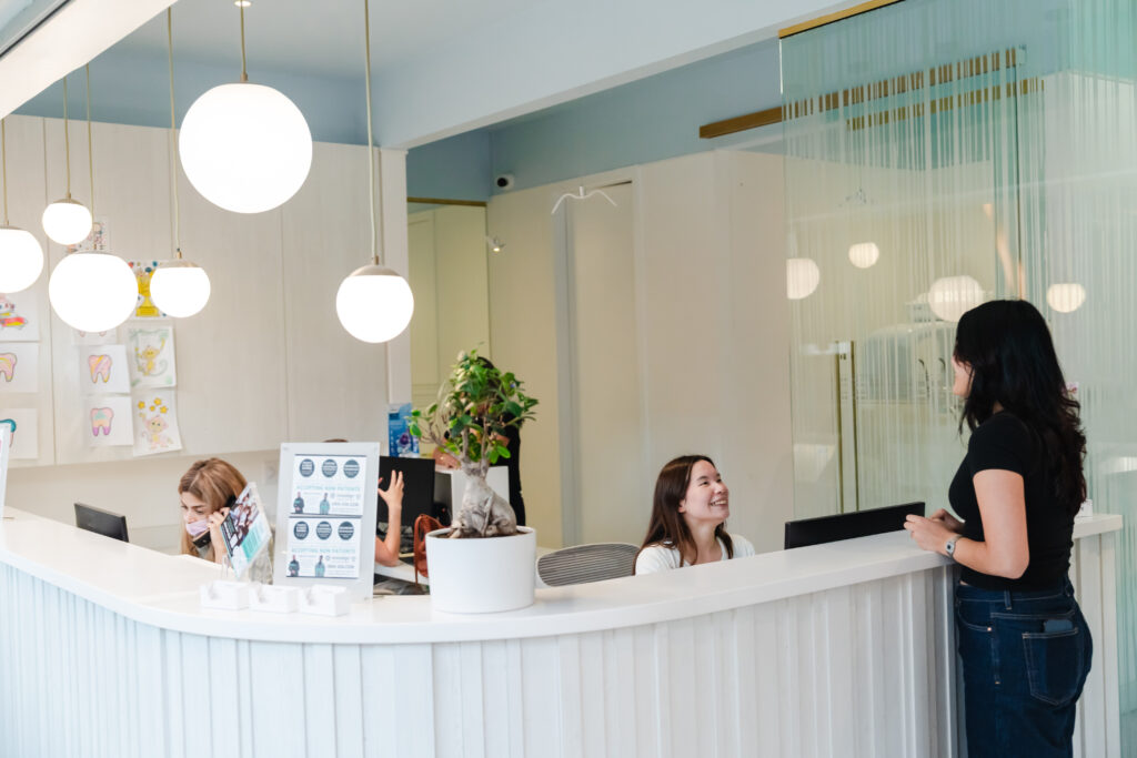 A friendly dental office reception area with two staff members engaged at the front desk while a visitor speaks with another staff member. The space is bright and welcoming, featuring modern lighting and greenery for a pleasant atmosphere. The 123Dentist Smiley is also present to add cheer.