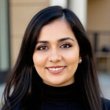 A smiling woman with dark hair and a black top stands outdoors, showcasing a warm and friendly expression against a blurred background.