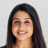 A smiling woman with long dark hair is posing against a light gray background, radiating warmth and positivity.