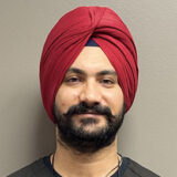 A smiling person wearing a red turban and dark attire is posing against a neutral gray background.